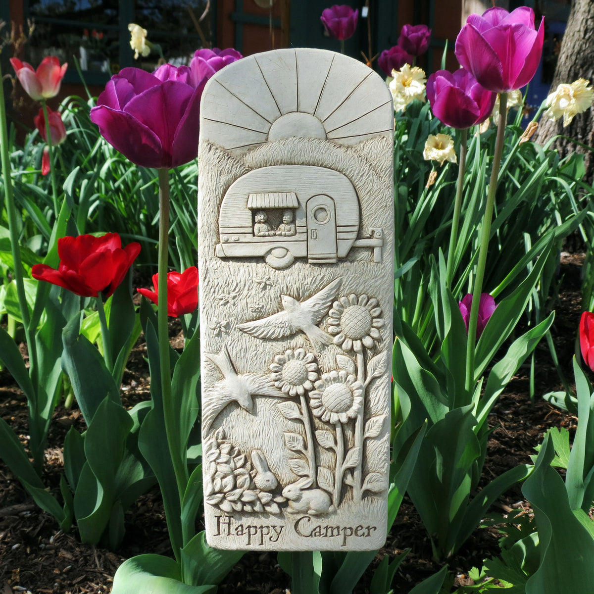 Happy Camper plaque standing in a spring garden bed among red and purple tulips